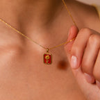A person holds the Alexandrite Birth Flower Charm Necklace, featuring a red and gold pendant with a tree design symbolizing growth and strength, close to their neck.