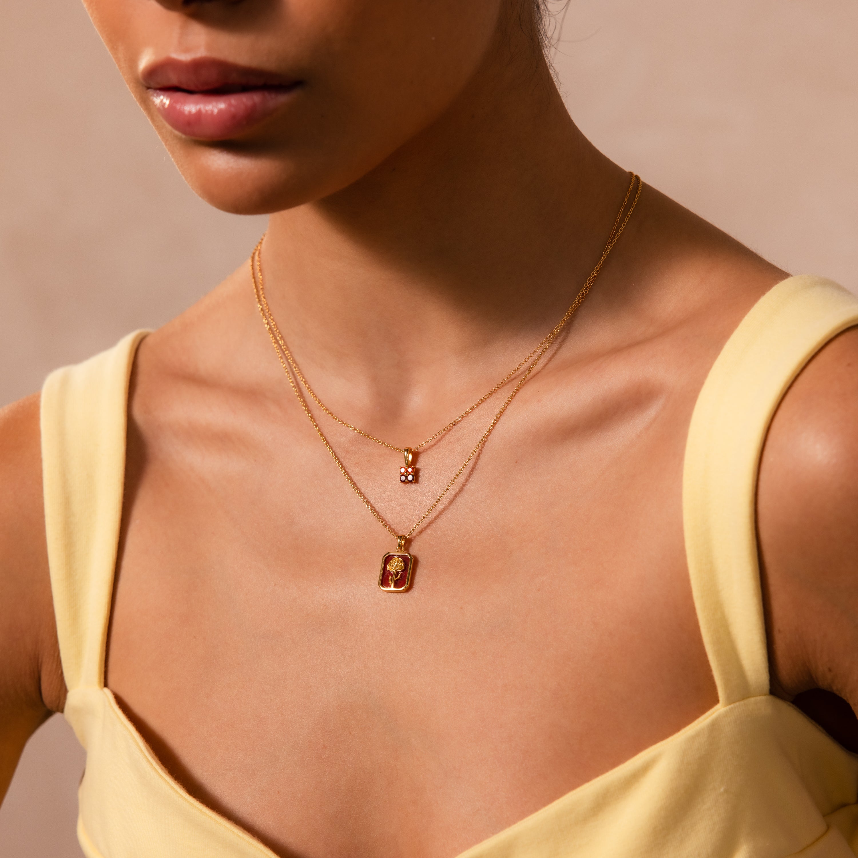 A woman in a yellow top wears two layered gold necklaces featuring small, rectangular Ruby Birth Flower Charm pendants.