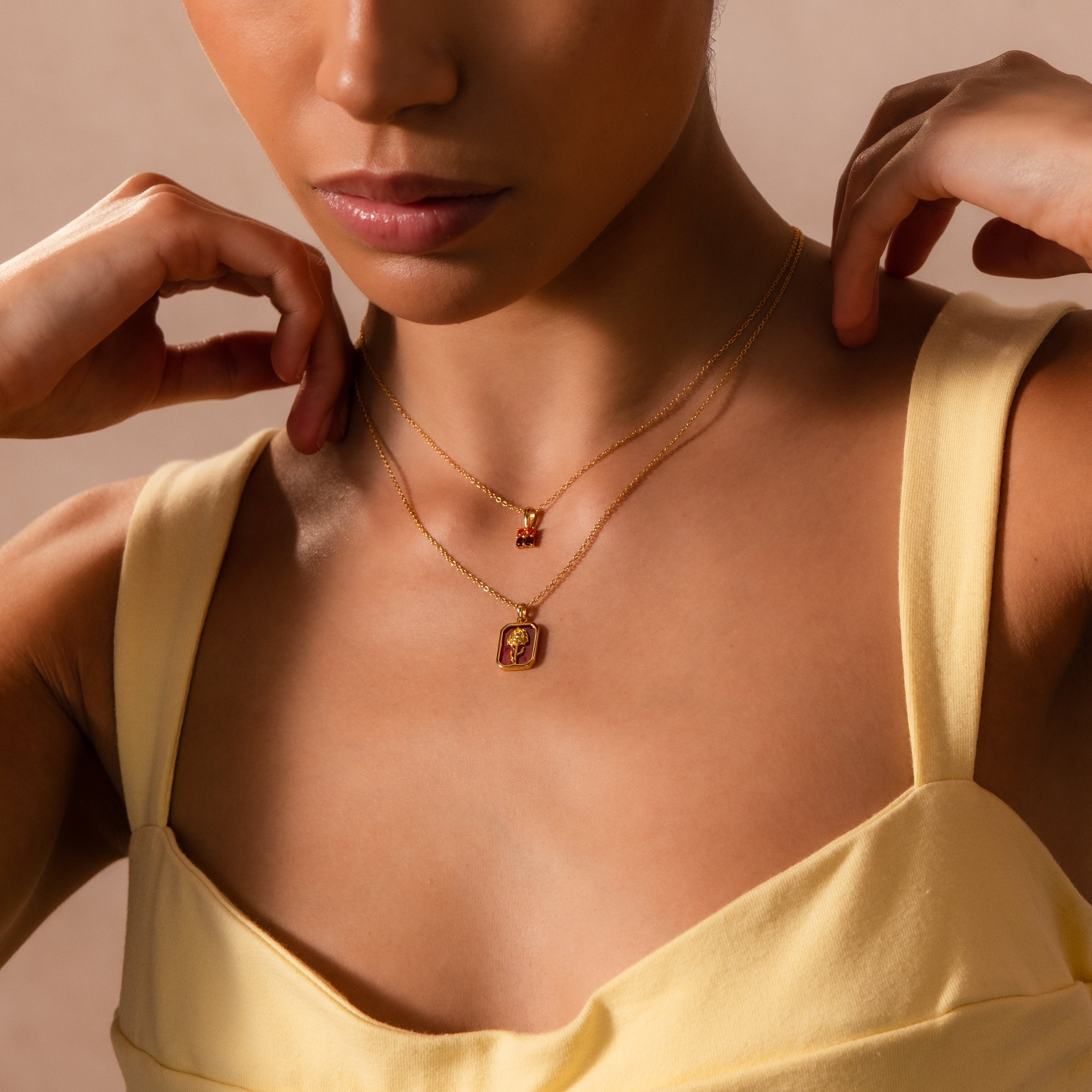 Woman in a yellow top adjusts her Garnet Birth Flower Charm Necklace, layered with another gold pendant.
