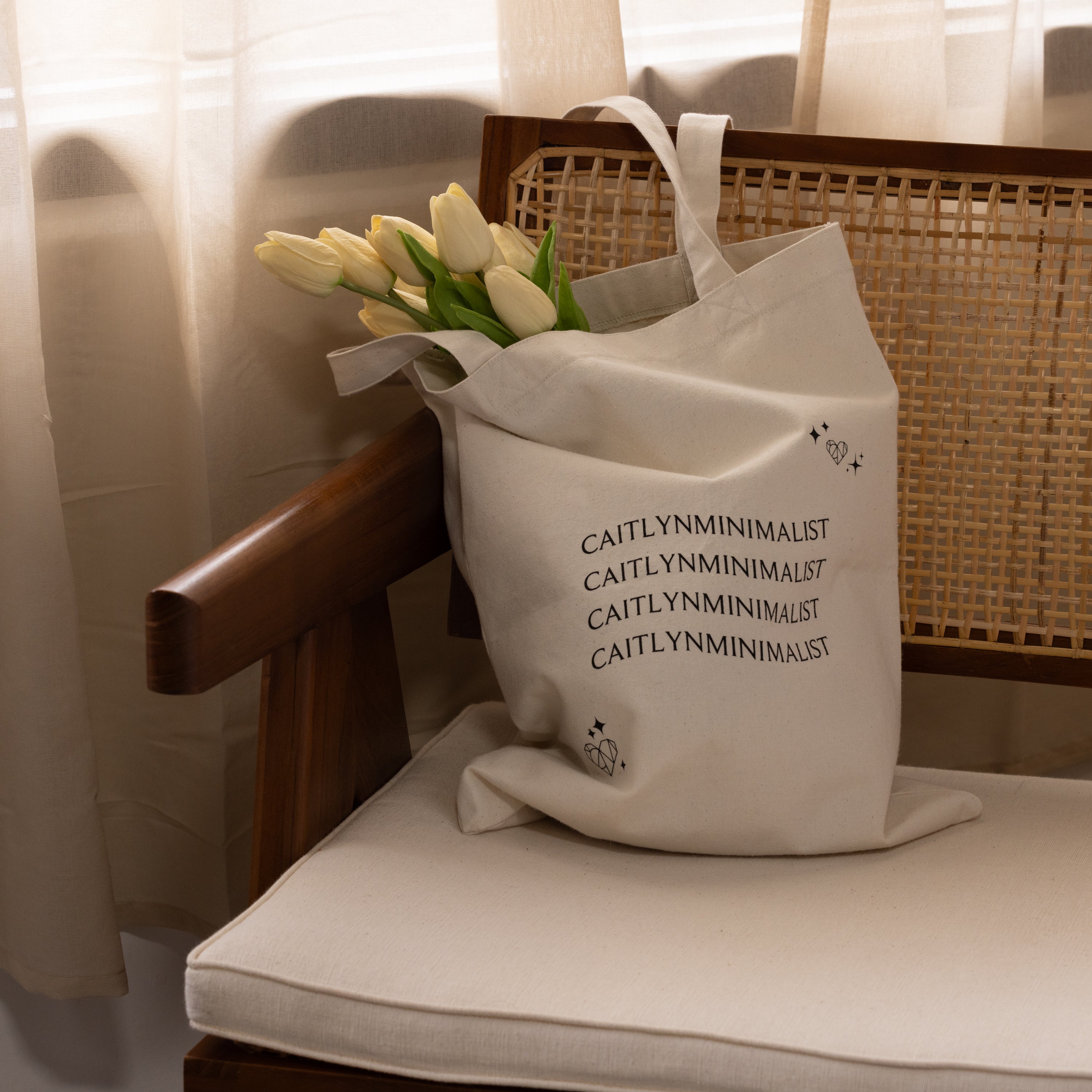 The CM Tote Bag in beige, featuring a minimalist design, is shown with tulips placed on a wooden chair as sunlight filters through sheer curtains in the background.