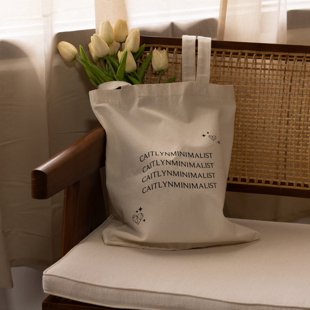 The CM Tote Bag in beige features white tulips and CAITLYNMINIMALIST text, styled on a wooden chair with a woven back.
