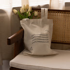 The CM Tote Bag in beige features white tulips and CAITLYNMINIMALIST text, styled on a wooden chair with a woven back.