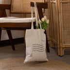 The CM Tote Bag, featuring chic text, hangs on a wooden chair and holds white tulips.