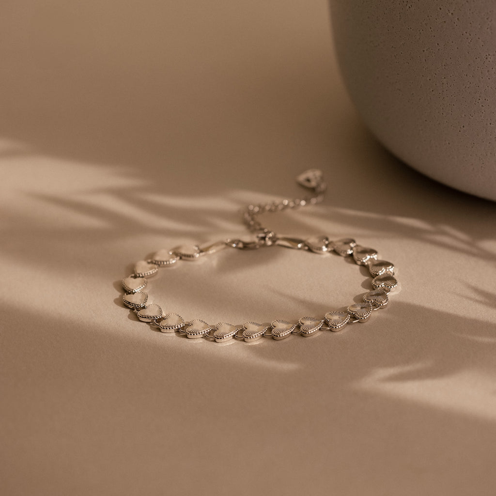 A product flat lay of the Ribbed Heart Link Bracelet in White Gold resting on a soft beige surface with diffused natural light. The bracelet forms a delicate curve, showing the spacing and articulation between the heart links. 
