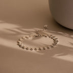 A product flat lay of the Ribbed Heart Link Bracelet in White Gold resting on a soft beige surface with diffused natural light. The bracelet forms a delicate curve, showing the spacing and articulation between the heart links. 