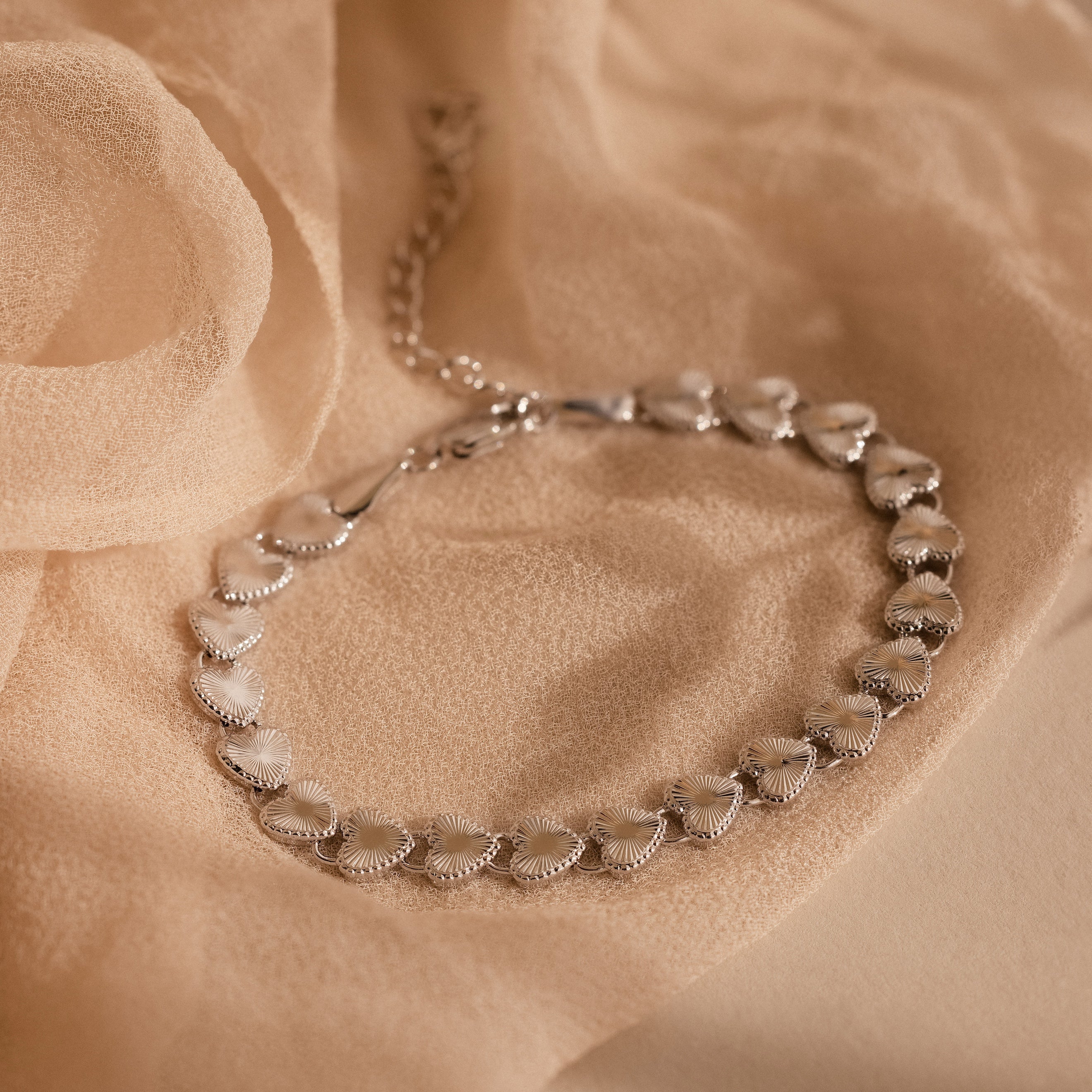 The Ribbed Heart Link Bracelet in White Gold is laid flat on a beige fabric backdrop, highlighting its textured, gleaming heart links and dainty chain clasp.