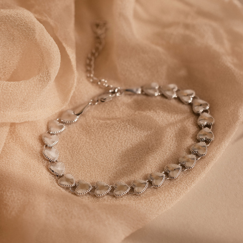 The Ribbed Heart Link Bracelet in White Gold is laid flat on a beige fabric backdrop, highlighting its textured, gleaming heart links and dainty chain clasp.