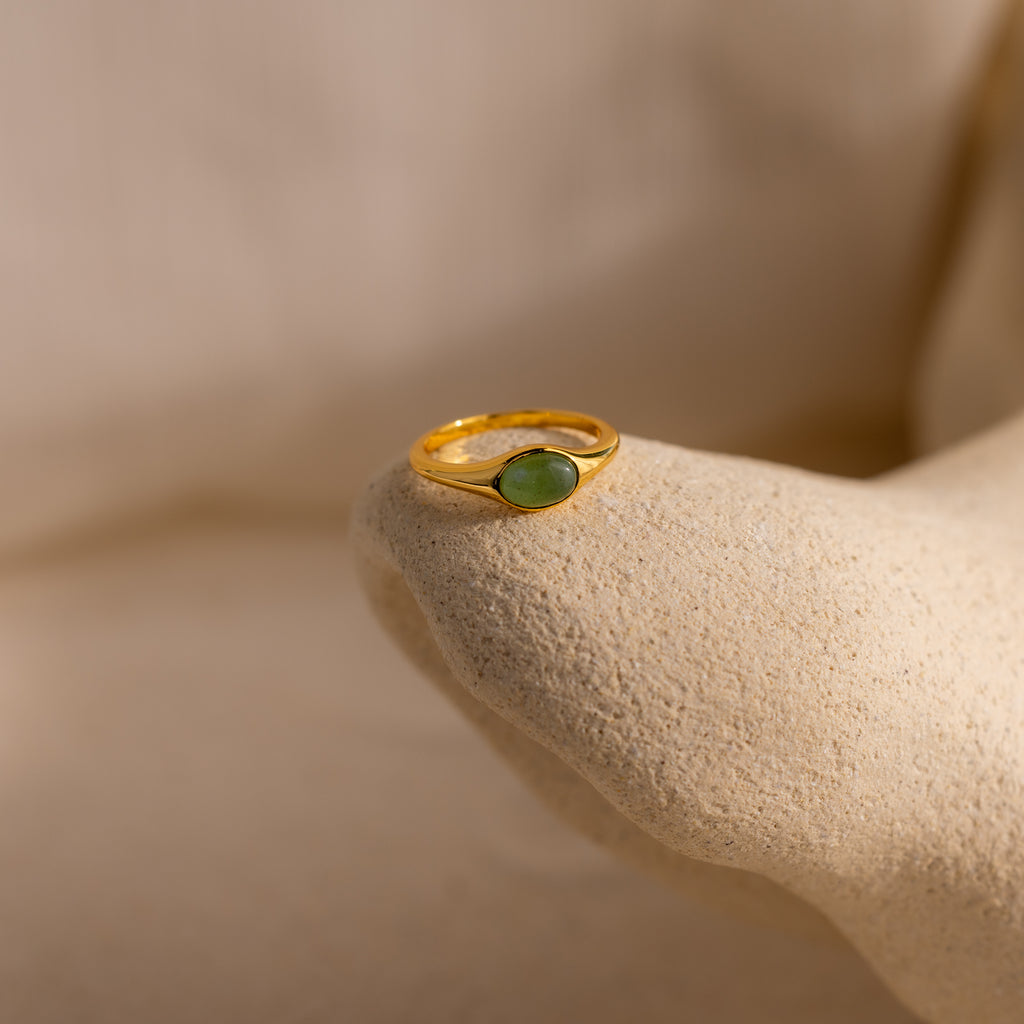 The Oval Jade Signet Ring with a green jade stone is displayed on a textured beige surface, highlighting its elegant design.