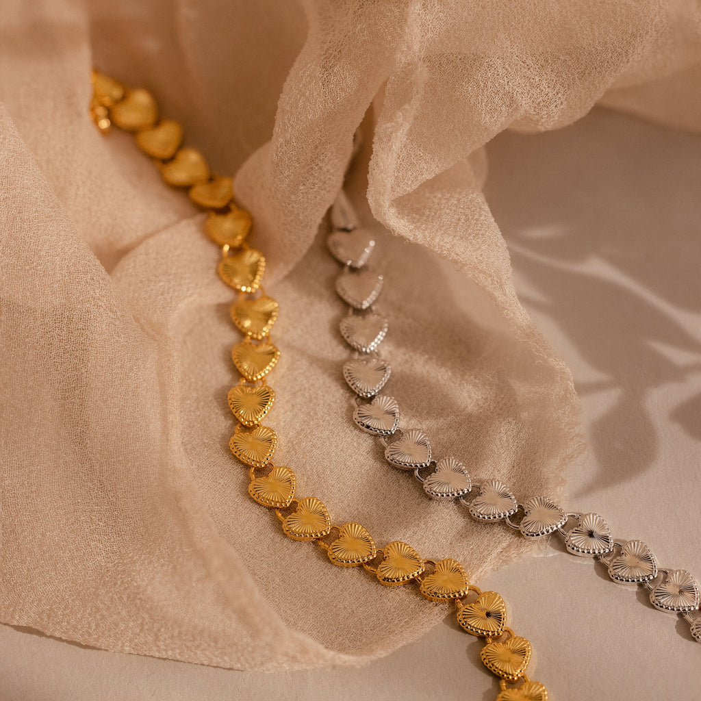 Two Ribbed Heart Link Bracelets, one in gold and one in white gold, are artfully arranged on soft beige fabric. Both display the ribbed heart pattern and fine craftsmanship.