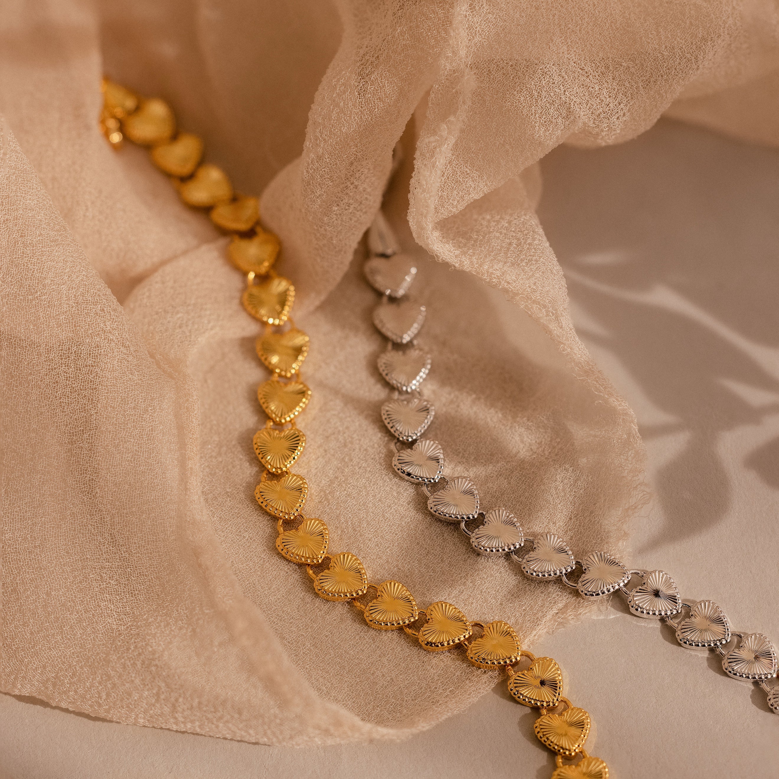 Two Ribbed Heart Link Bracelets, one in gold and one in white gold, are artfully arranged on soft beige fabric. Both display the ribbed heart pattern and fine craftsmanship.