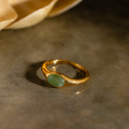 The Oval Jade Signet Ring features a vibrant green jade stone set in gold, displayed on a gray surface with a soft beige background.