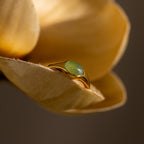 The Oval Jade Signet Ring features a green gem set on a beige flower petal, softly lit with a blurred background—an exquisite piece of elegant jewelry.