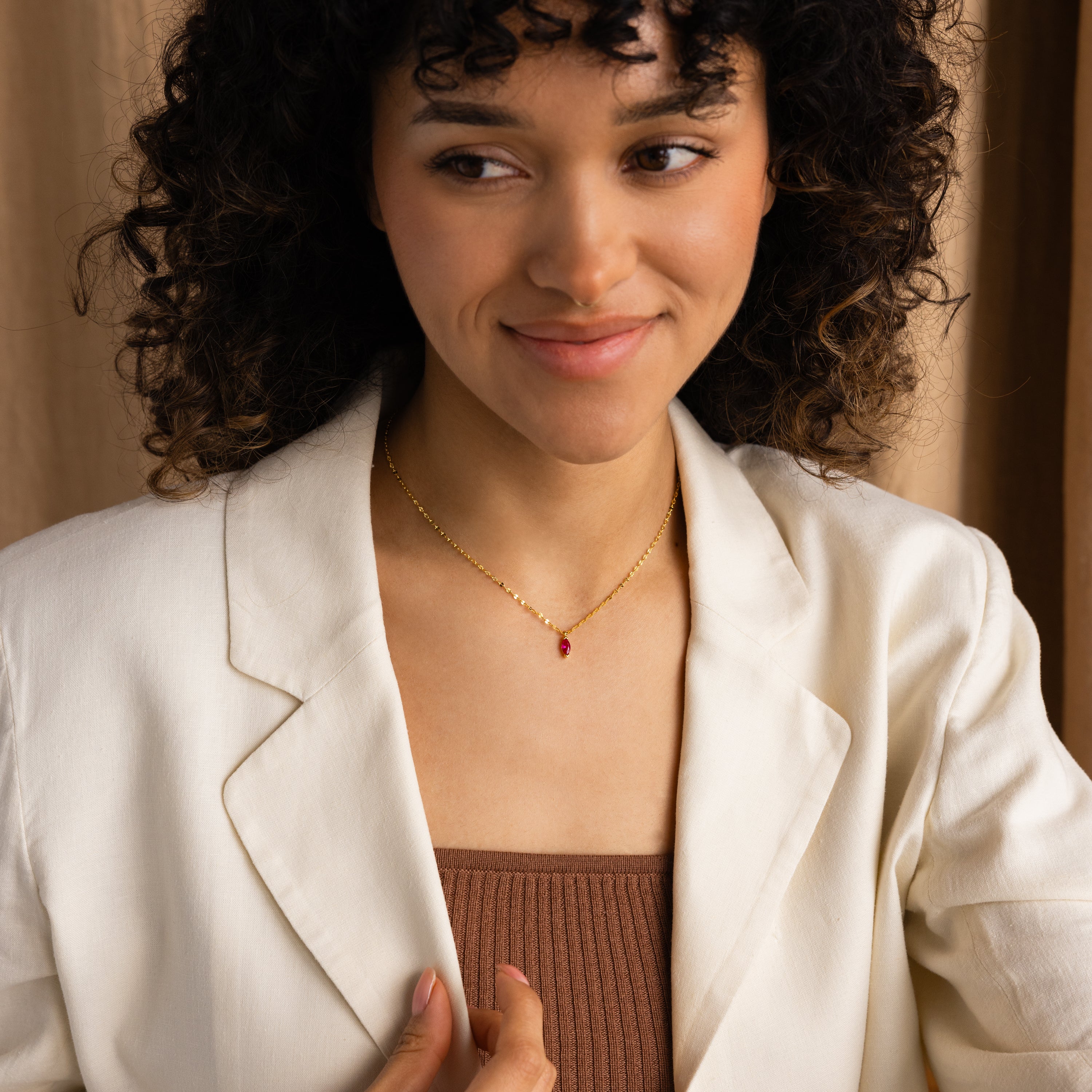 A woman in a white blazer and brown top smiles softly, wearing the Custom Marquise Birthstone Necklace featuring a small red marquise-cut pendant.