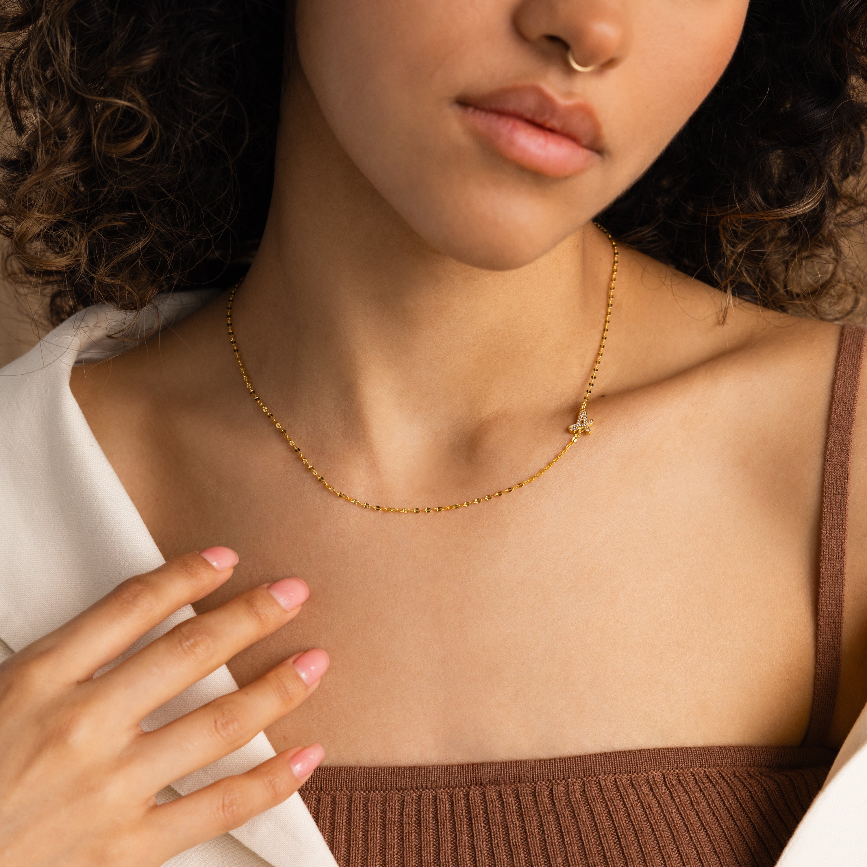 A woman wears the Nella Pave Off-Center Initial Necklace with a white shirt, brown top, manicured nails, and a nose ring.
