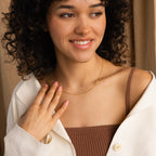 Smiling woman with curly hair wearing a cream jacket over a brown top and the Nella Pave Off-Center Initial Necklace.