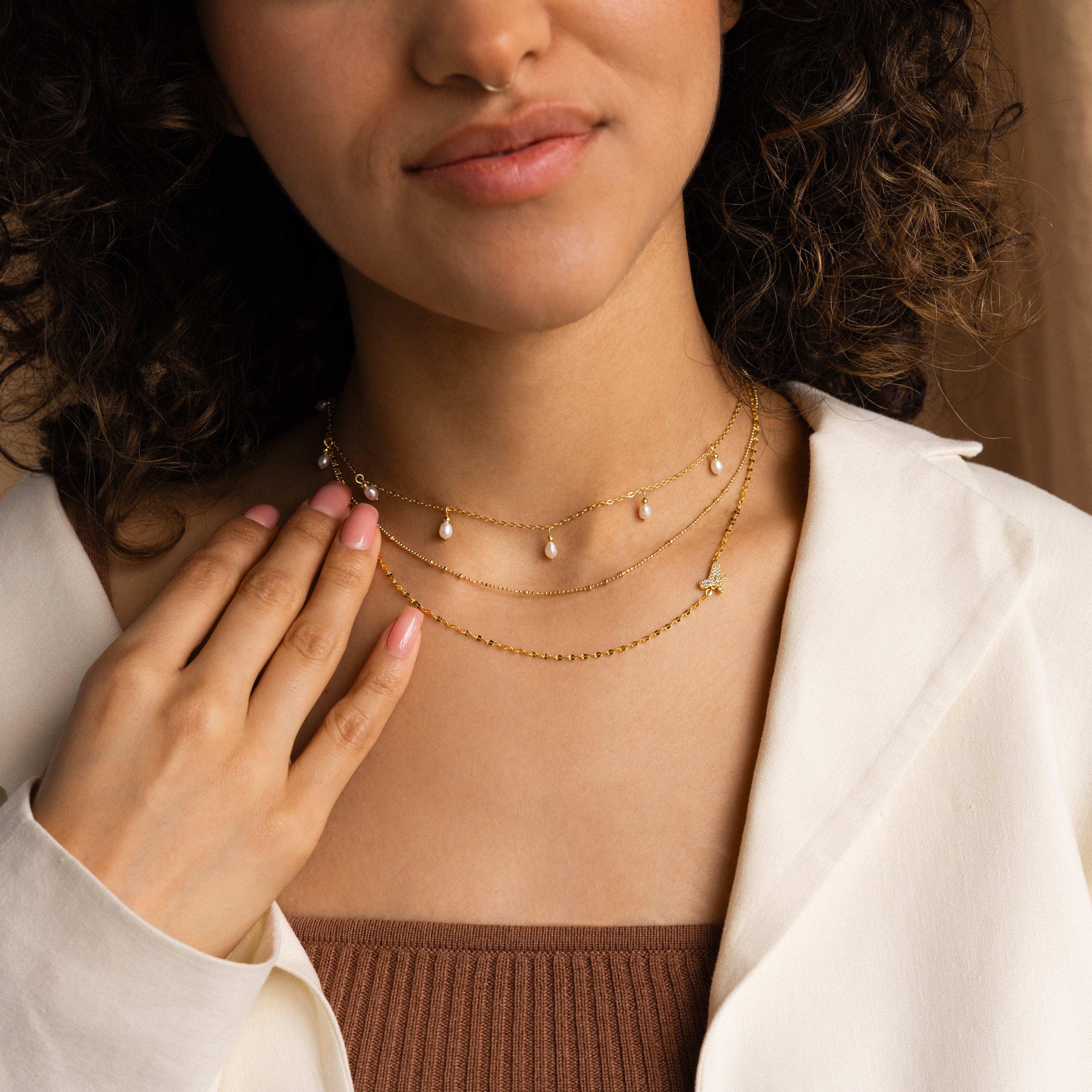 Woman wearing layered gold necklaces, featuring the Nella Pave Off-Center Initial Necklace, styled with a white blazer and her hand gently touching her collarbone.