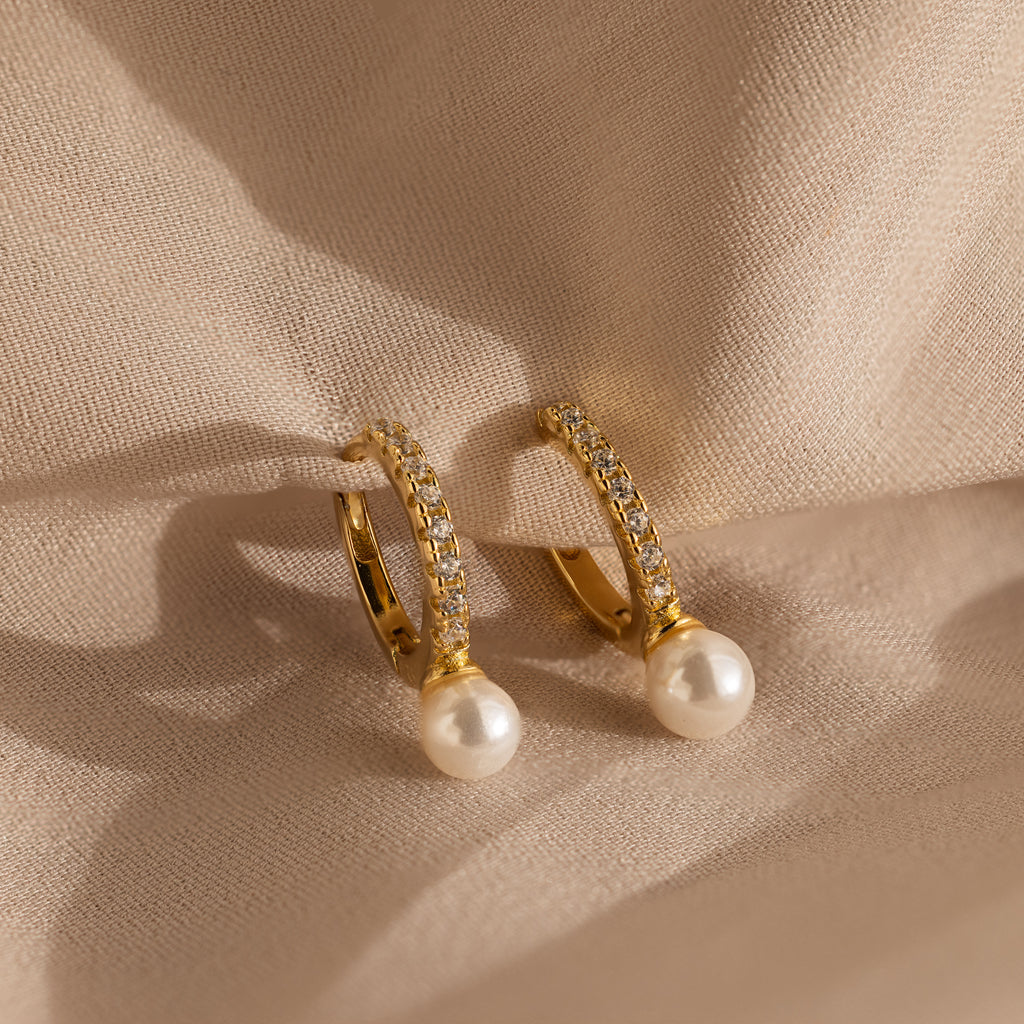 The Pave Pearl Drop Huggies, featuring gold hoops with pearls and small diamonds, are elegantly displayed on soft beige fabric.