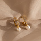 The Pave Pearl Drop Huggies, featuring gold hoops with pearls and small diamonds, are elegantly displayed on soft beige fabric.