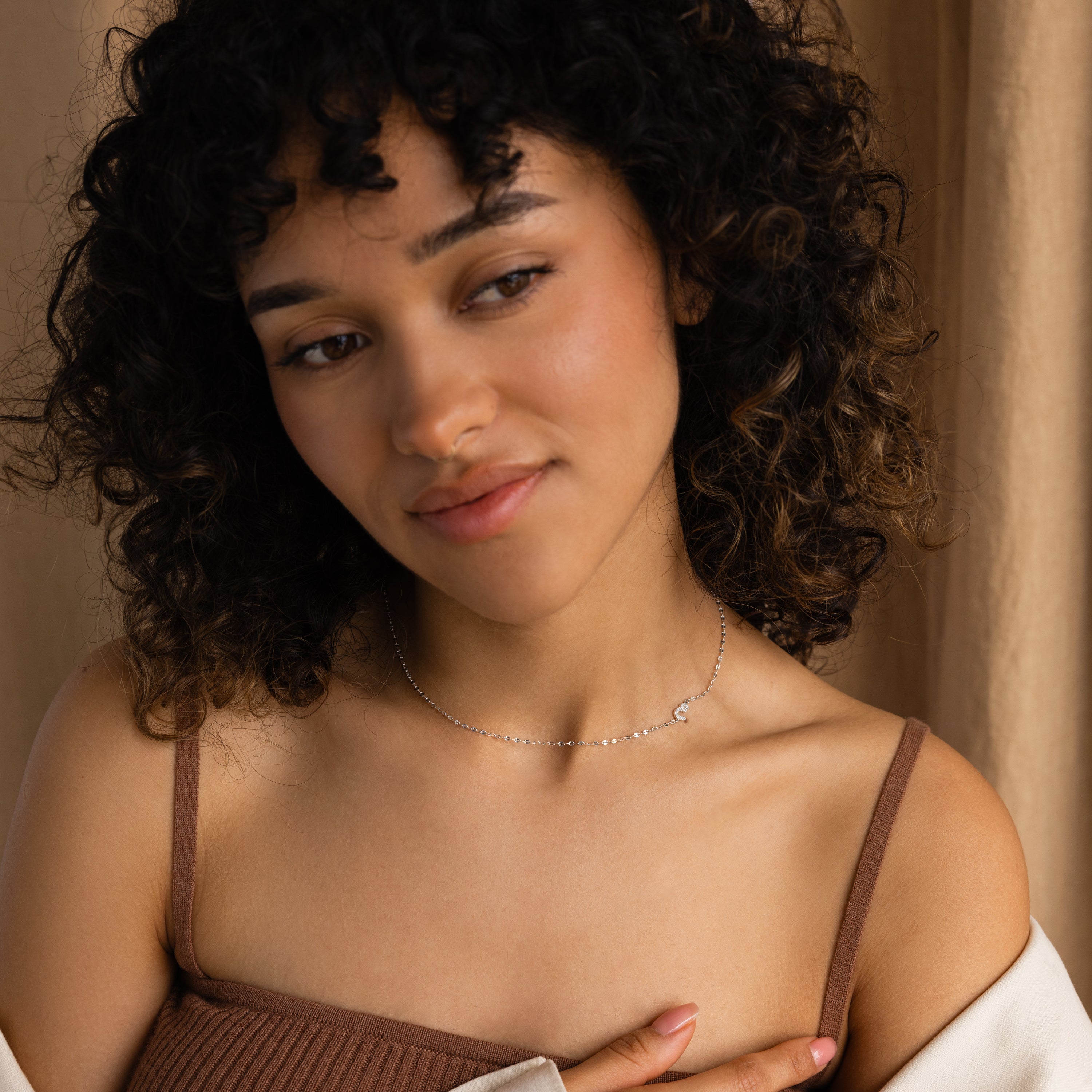 A woman with curly hair, wearing a brown top and the Nella Pave Off-Center Initial Necklace, looks to the side with a gentle smile.