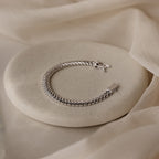 The V-Link Chain Tennis Bracelet, featuring sculptural silver links, is showcased on a round light stone with a soft beige fabric background.