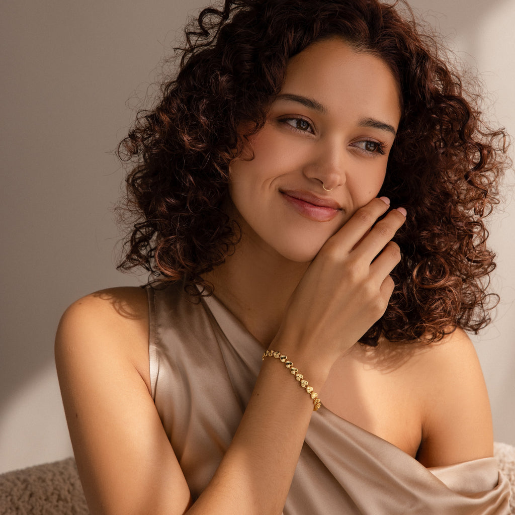 A smiling woman with curly hair wearing the Ribbed Heart Charm bracelet, her hand gently touching her face.
