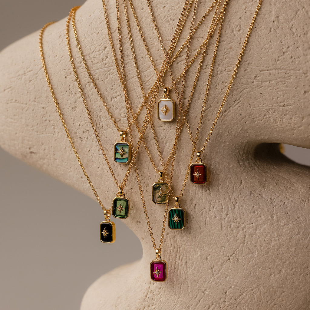 The North Star Ruby Necklace features a gold chain and a colorful rectangular pendant with a sparkling diamond-set North Star, elegantly displayed on a textured beige stand.