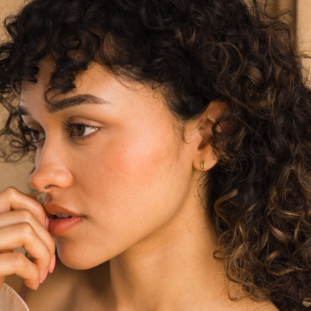 A woman with curly hair and gold gemstone jewelry gazes thoughtfully, her hand near her lips, highlighting the elegant Tapered Baguette Birthstone Studs.