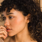A woman with curly hair and gold gemstone jewelry gazes thoughtfully, her hand near her lips, highlighting the elegant Tapered Baguette Birthstone Studs.