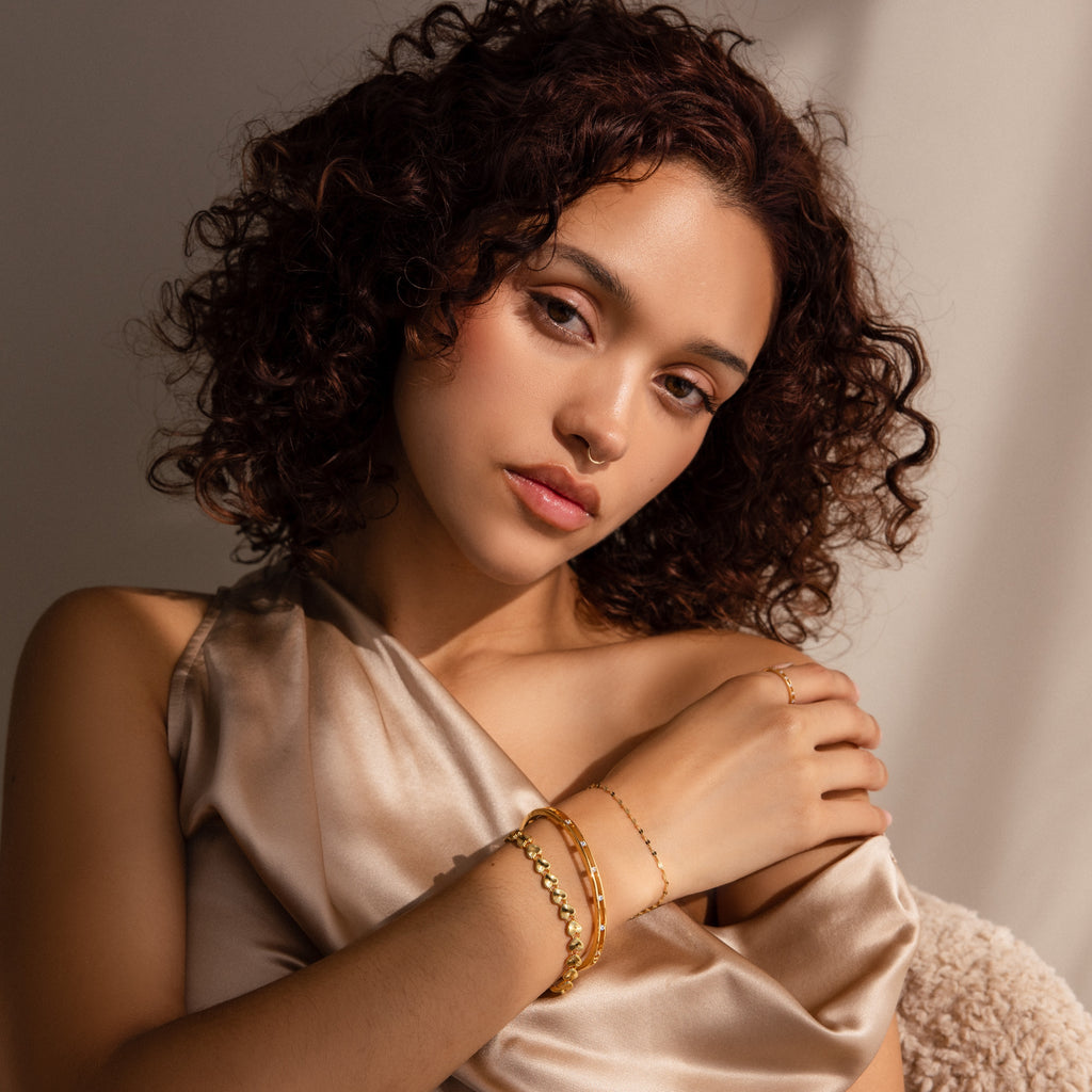 Model wears The Ribbed Heart Link Bracelet,  stacked with two other bracelets: a thick gold bangle with inset stones and a fine, delicate gold chain. 