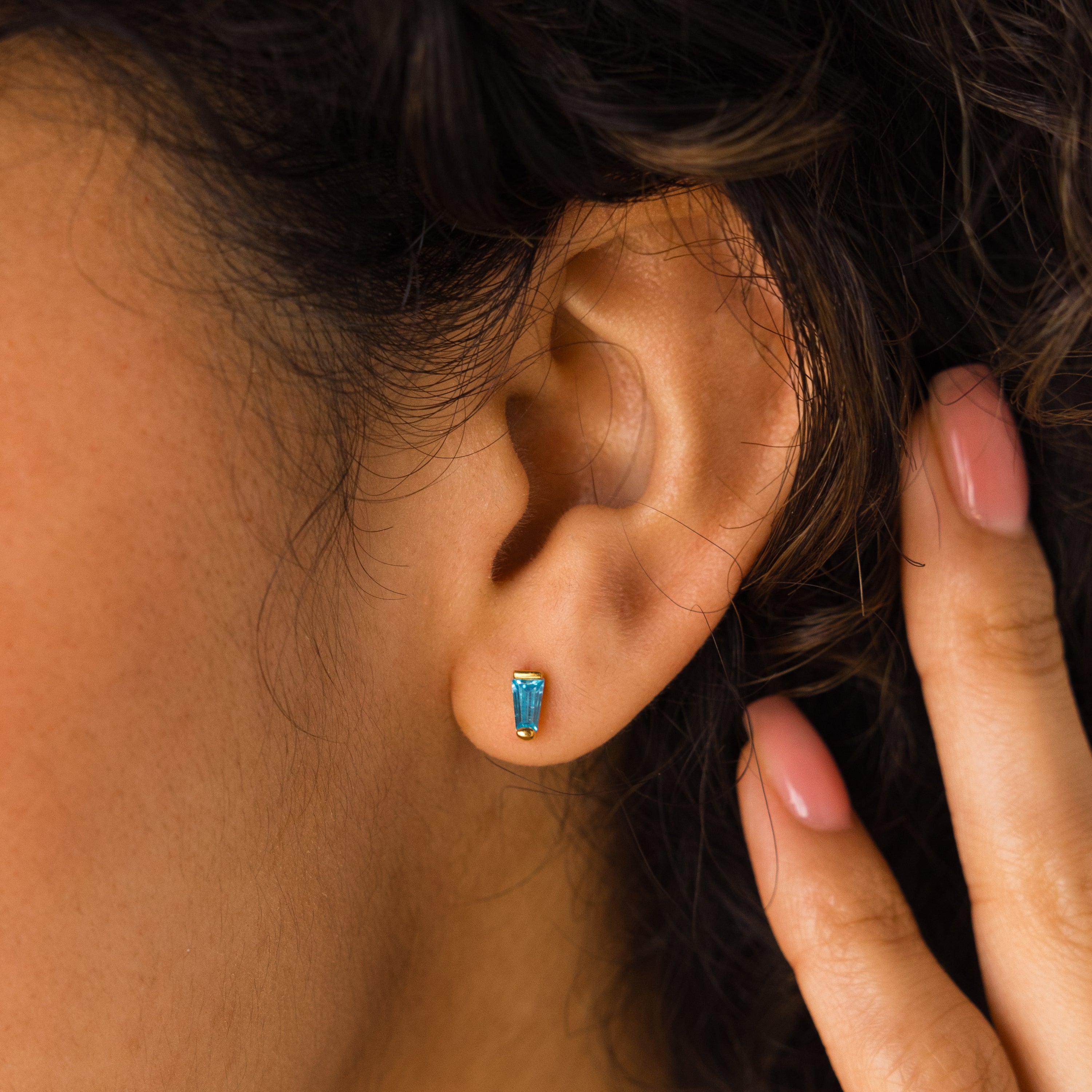 Close-up of a woman's ear adorned with a small blue Tapered Baguette Birthstone Studs earring, her hand gently near her ear—a delicate gemstone jewelry piece.