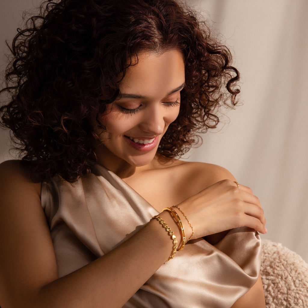 A smiling woman with curly hair wears multiple gold bracelets, including a version of the Ribbed Heart Link Bracelet in gold.