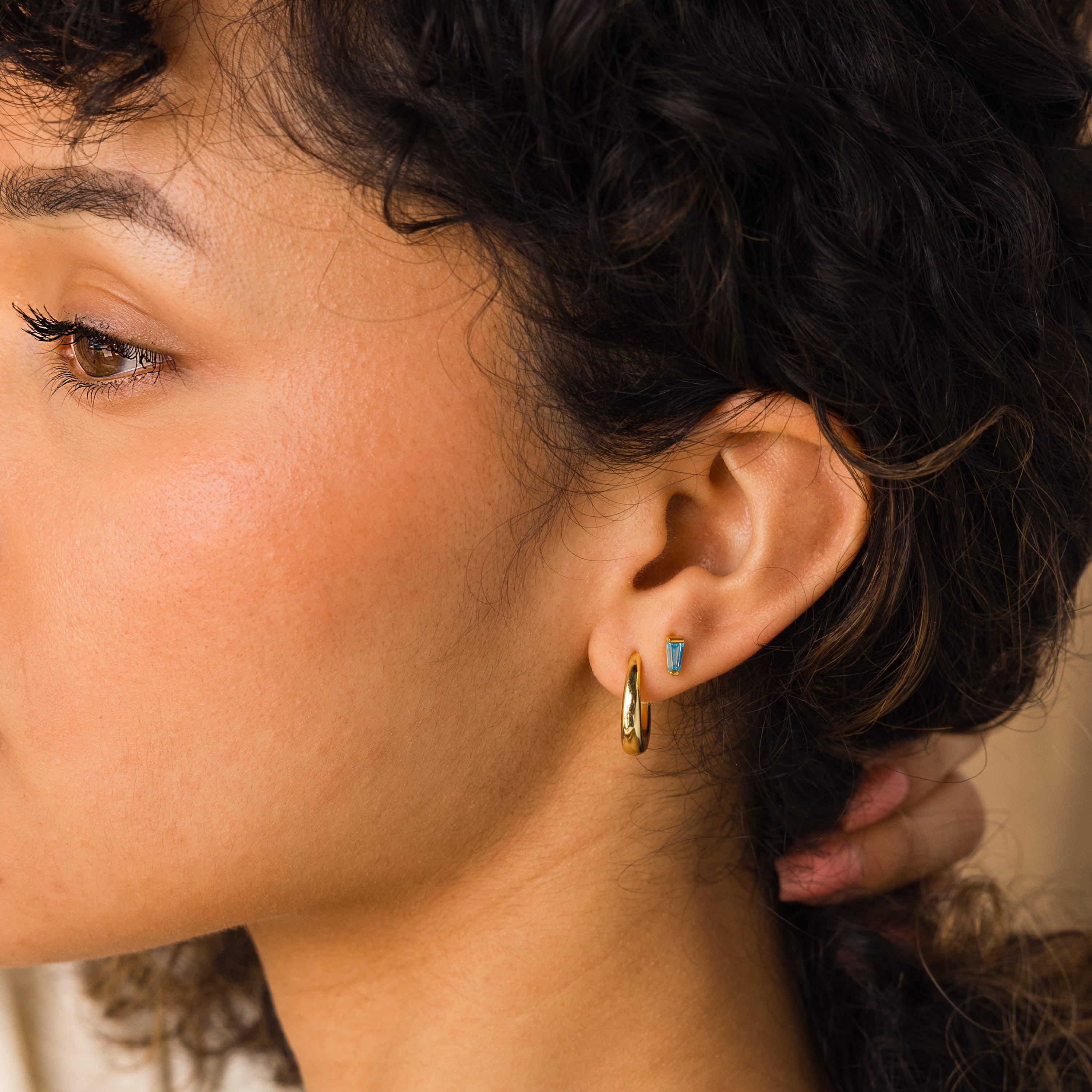 Close-up of a woman’s ear wearing Tapered Baguette Birthstone Studs and a gold hoop, with curly hair visible—ideal for gemstone jewelry lovers.