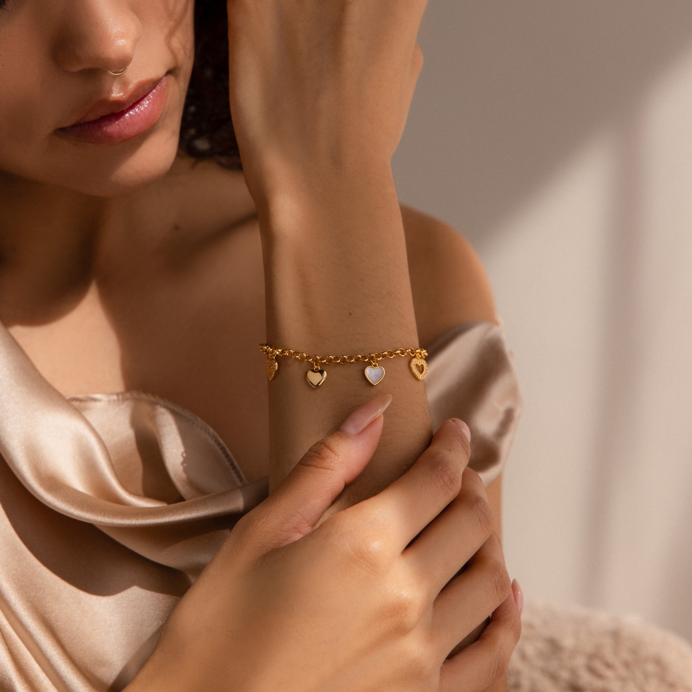 A woman wears the Heart Charm Bracelet with gold heart charms, paired with a silky beige off-shoulder top—an ideal gift for your girlfriend.