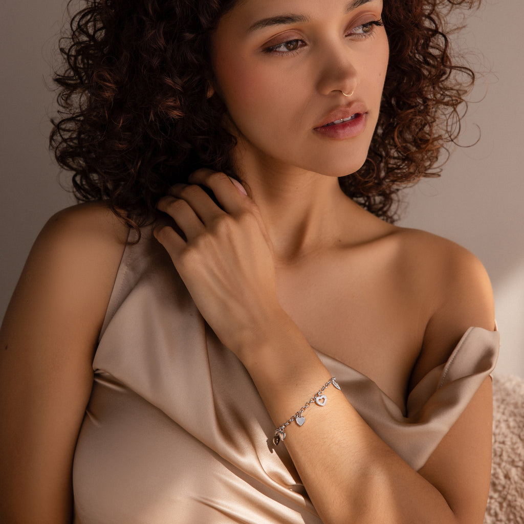 A woman with curly hair wears a satin off-shoulder top and the Heart Charm Bracelet, gazing thoughtfully to the side—a stylish gift for any girlfriend who loves elegant heart-themed jewelry.