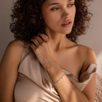 A woman with curly hair wears a satin off-shoulder top and the Heart Charm Bracelet, gazing thoughtfully to the side—a stylish gift for any girlfriend who loves elegant heart-themed jewelry.