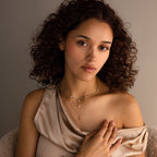 A woman with curly hair wears the Ivy Diamond Station Necklace, featuring diamond accents, layered with gold necklaces and a beige one-shoulder top, looking at the camera.