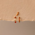 The Garnet Earring Trio Set—three gold earrings with red gemstones, including a huggie hoop—are arranged on torn beige paper for a striking display.
