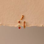 The Garnet Earring Trio Set—three gold earrings with red gemstones, including a huggie hoop—are arranged on torn beige paper for a striking display.