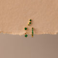 The Emerald Earring Trio Set features gold earrings and studs with green gemstones, including an elegant birthstone pair and pavé huggie hoops, displayed on a textured beige surface.