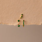 The Emerald Earring Trio Set features gold earrings and studs with green gemstones, including an elegant birthstone pair and pavé huggie hoops, displayed on a textured beige surface.
