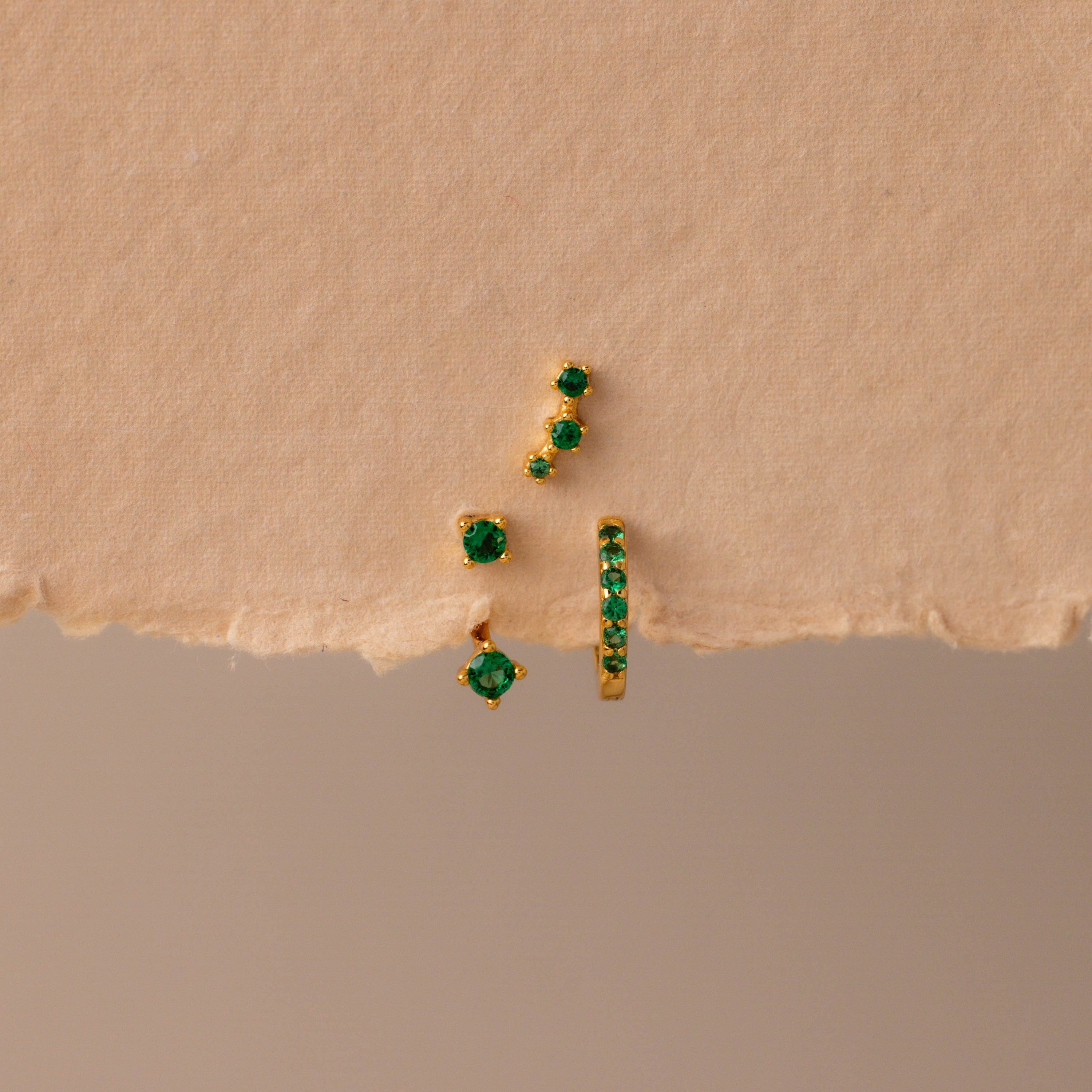 The Emerald Earring Trio Set features gold earrings and studs with green gemstones, including an elegant birthstone pair and pavé huggie hoops, displayed on a textured beige surface.