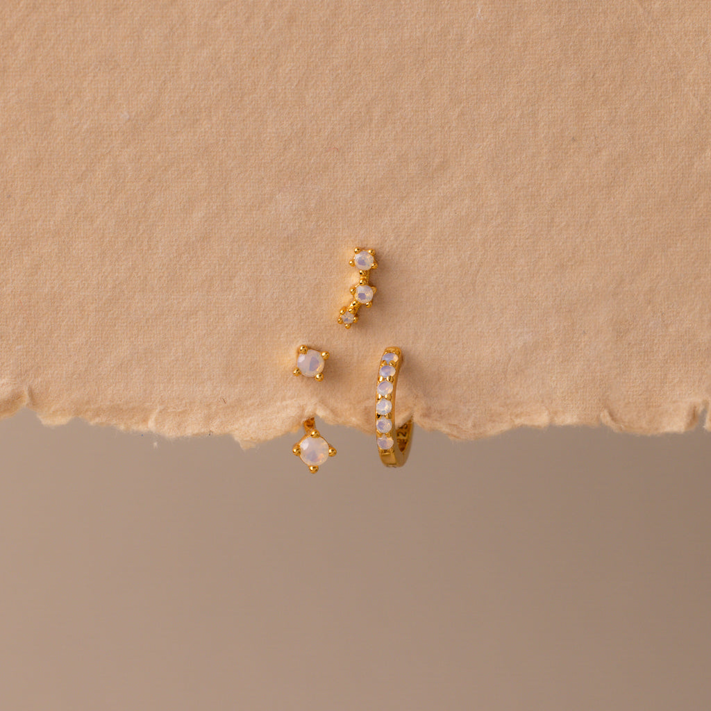 The Opal Earring Trio Set, featuring three delicate gold and pearl earrings including a huggie hoop style, is artfully arranged on a textured beige background.
