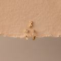 The Opal Earring Trio Set, featuring three delicate gold and pearl earrings including a huggie hoop style, is artfully arranged on a textured beige background.