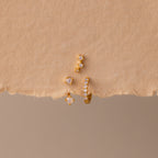 The Opal Earring Trio Set, featuring three delicate gold and pearl earrings including a huggie hoop style, is artfully arranged on a textured beige background.