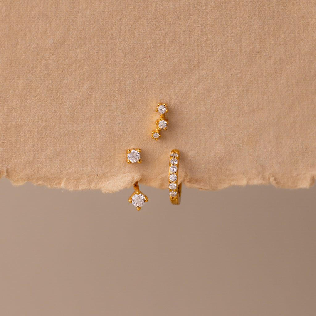The Diamond Earring Trio Set sparkles elegantly with small white gemstones on a textured beige surface, ideal for anyone who loves versatile birthstone earrings.