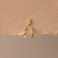 The Diamond Earring Trio Set sparkles elegantly with small white gemstones on a textured beige surface, ideal for anyone who loves versatile birthstone earrings.
