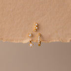 The Diamond Earring Trio Set sparkles elegantly with small white gemstones on a textured beige surface, ideal for anyone who loves versatile birthstone earrings.