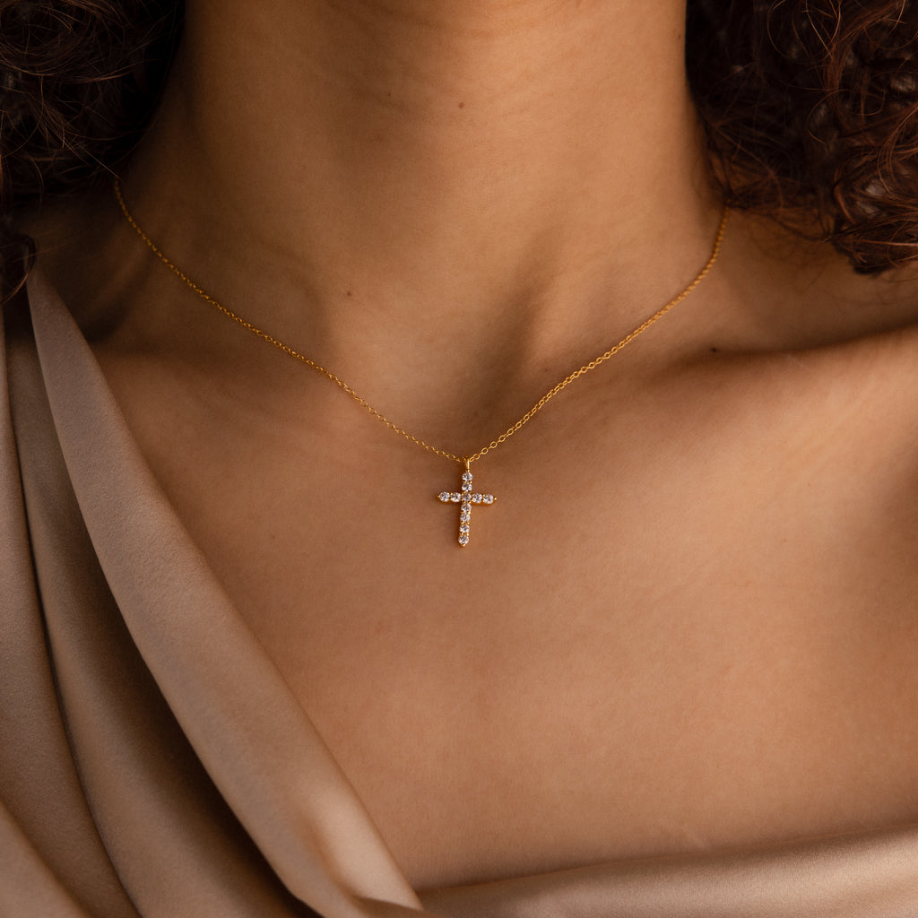 A woman wears the Pave Cross Necklace, featuring a sparkling jeweled pendant, elegantly styled with a beige off-shoulder blouse.