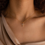 A woman wears the Pave Cross Necklace, featuring a sparkling jeweled pendant, elegantly styled with a beige off-shoulder blouse.