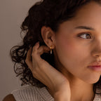 A woman with curly hair wears Dangling Pearl Diamond Huggies in 18K Gold and a white knit top, gently touching her ear and looking thoughtful.